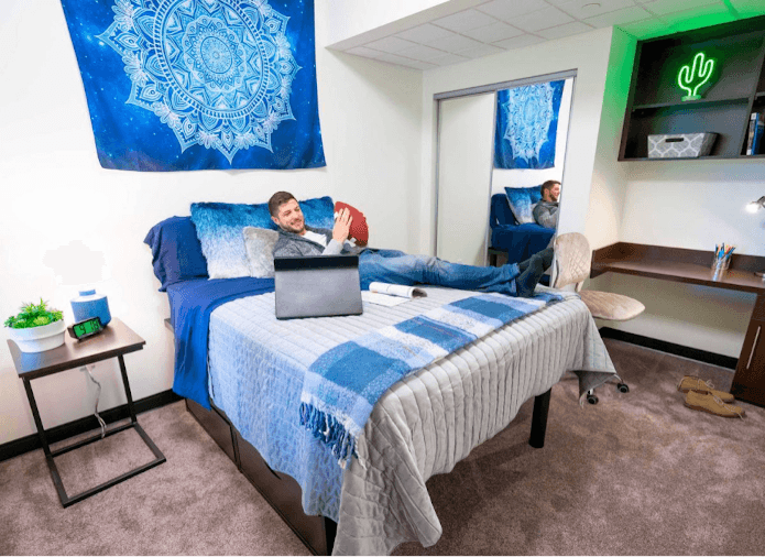 A Staged dorm room with white and blue sheets and a person on the bed with a laptop