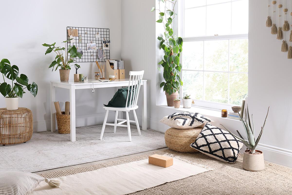 A light colored corner of a room with throw pillows on the floor, a desk and chair, accented by potted green house plants.