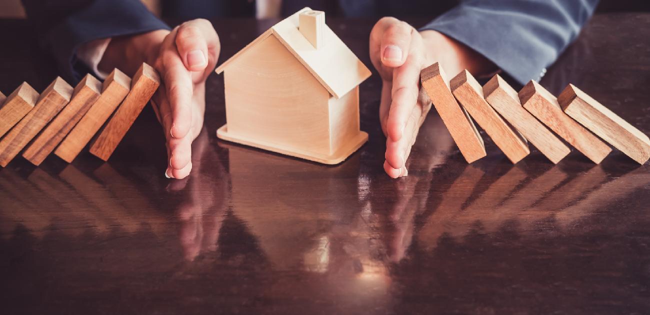 A wooden house miniature protected on both sides from falling dominoes by a pair of hands.