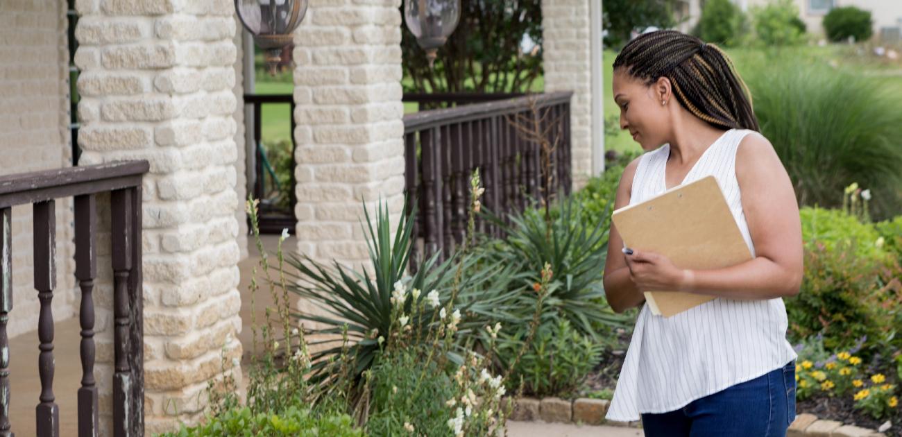 A woman stands outside a home, holding a clipboard and looking down at green plants grown in front of the home's porch.