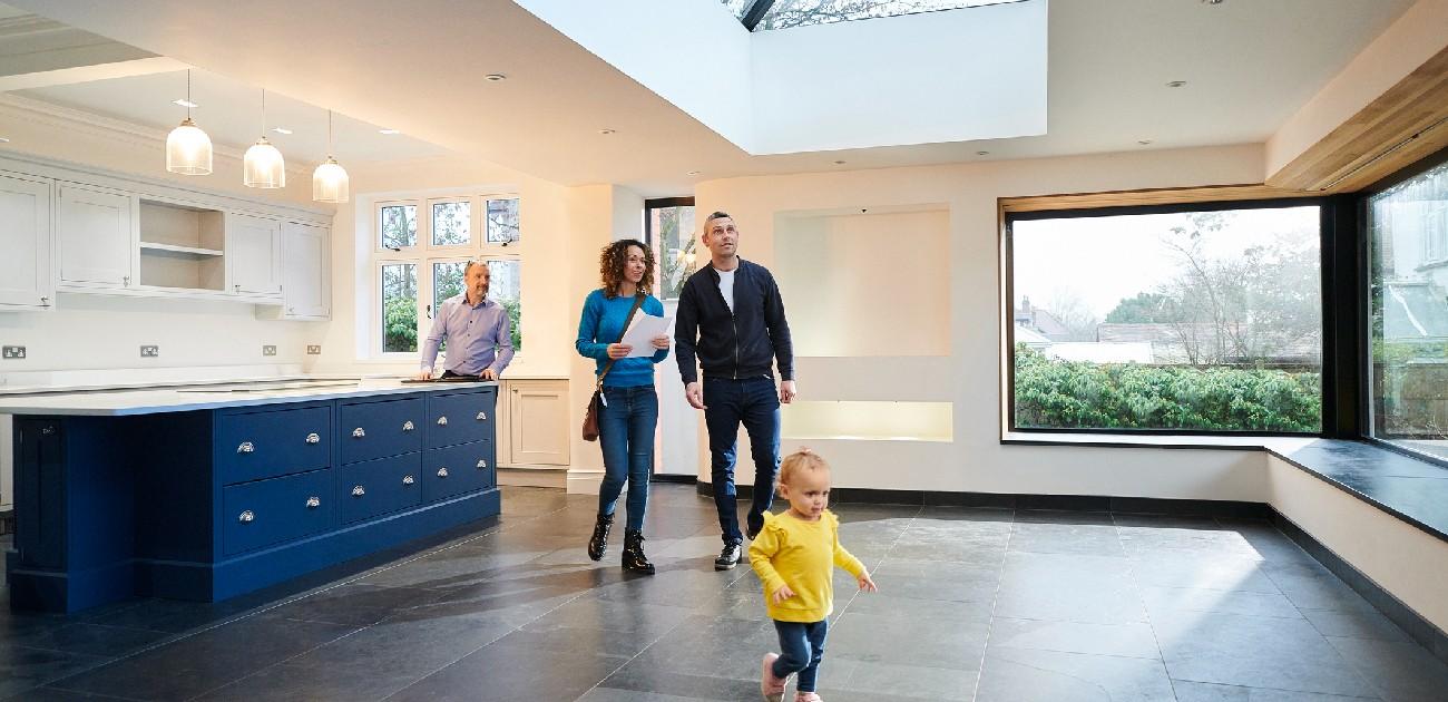 A man and woman stand in an open-format kitchen in a home, with a toddler running playfully in front of them.