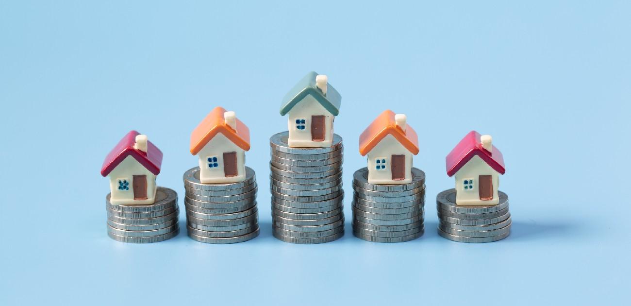 A picture of five small house miniatures each resting upon a stack of coins varying in height.