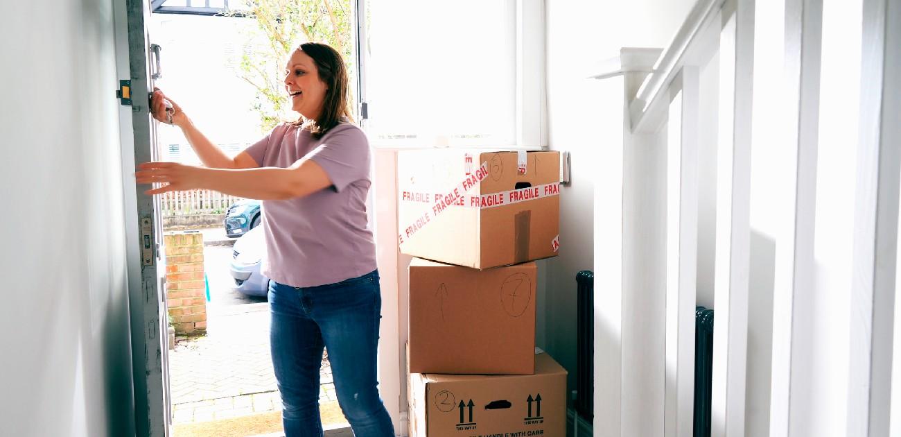 A woman stands at the front door on the inside of a new home, with moving boxes behind her next to a staircase leading up.