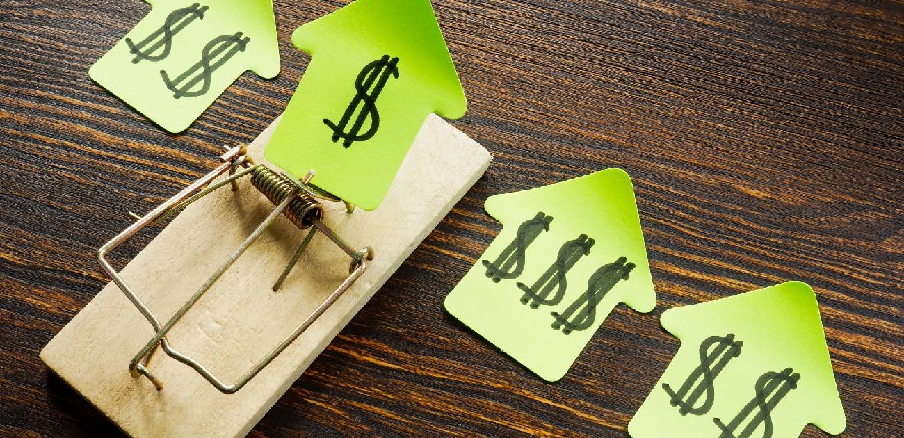 Picture of a mouse trap holding a paper cutout in the shape of a house with dollar signs written on it next to other cutouts.