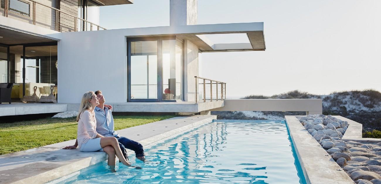 A man and a woman sit with their feet in the water of the pool outside their luxury home.