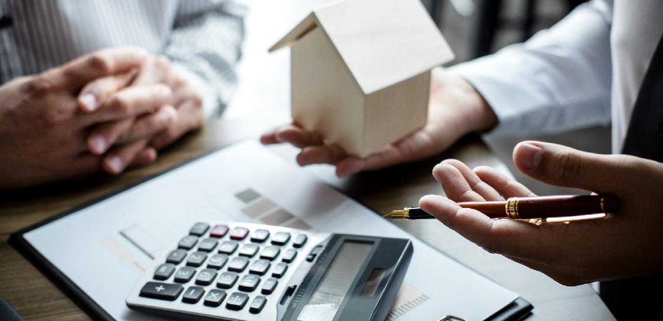 Two people sit at a desk going over a budget, with a calculator and house miniature in frame.