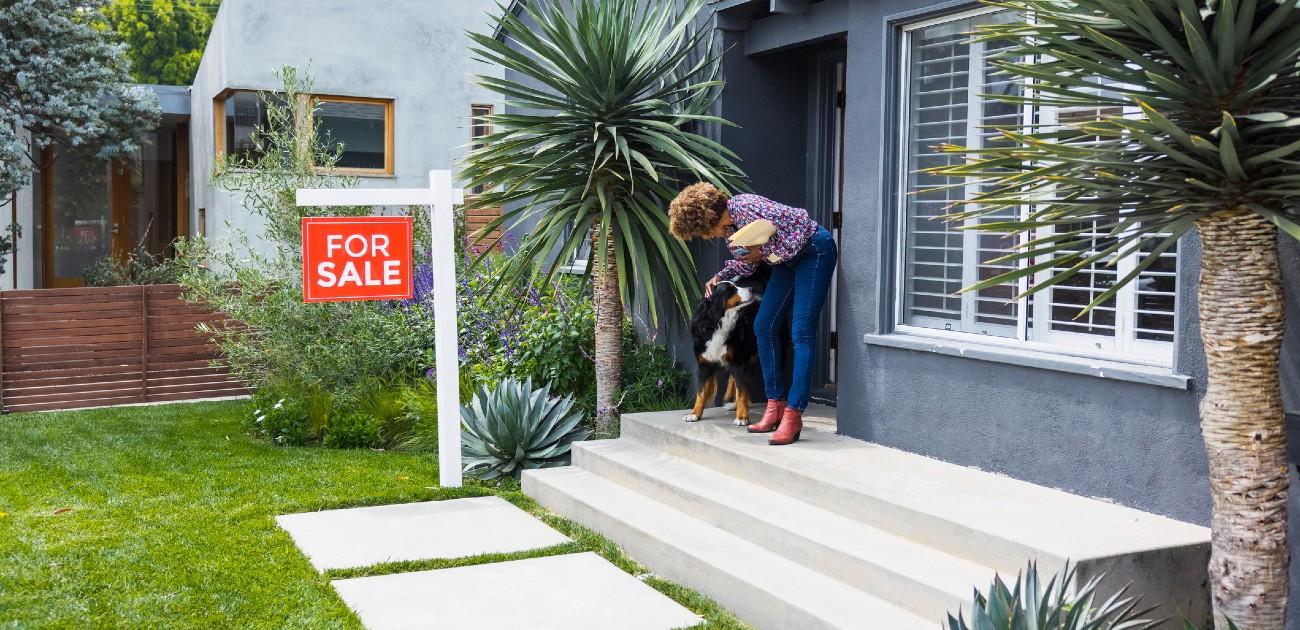 A woman stands on her front porch with her dog, with a "For Sale" sign a few feet ahead of her in the yard.