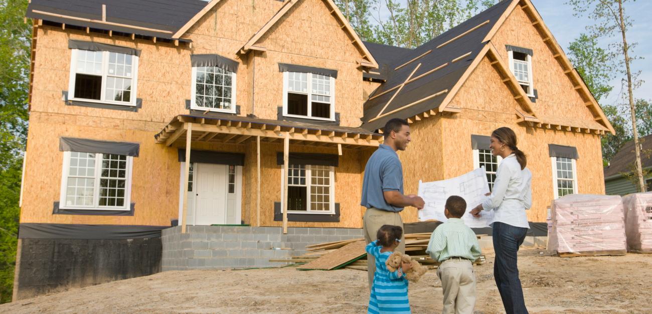 A family of four examines the plans for their partially constructed new home, which stands behind them.