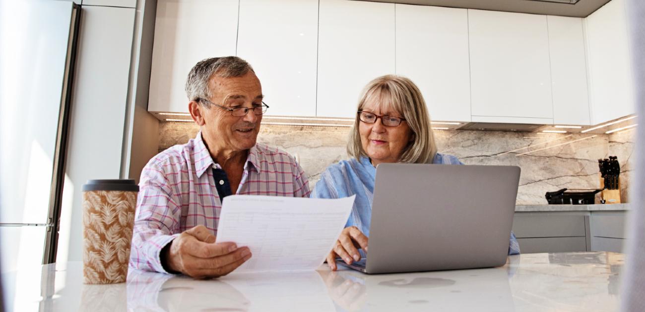 Married couple in their 60s, sitting at a kitchen table reviewing paperwork. Both are dressed casually.