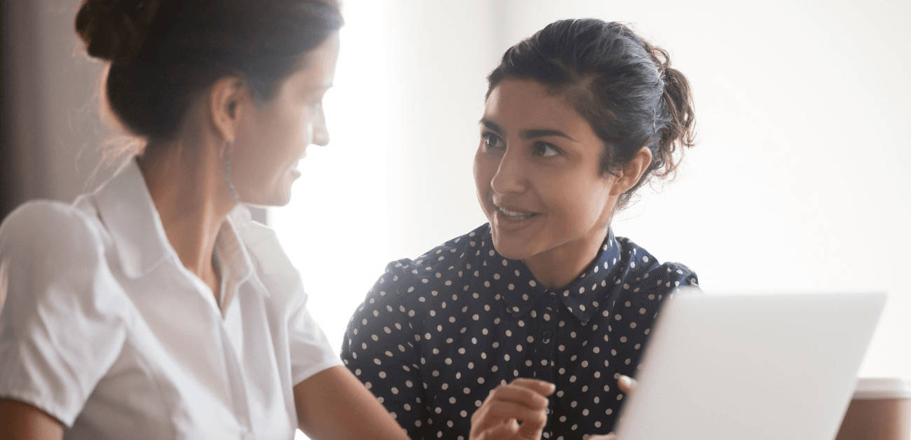 A woman with dark brown hair sitting in front of a laptop and talking with another woman, also with brown hair