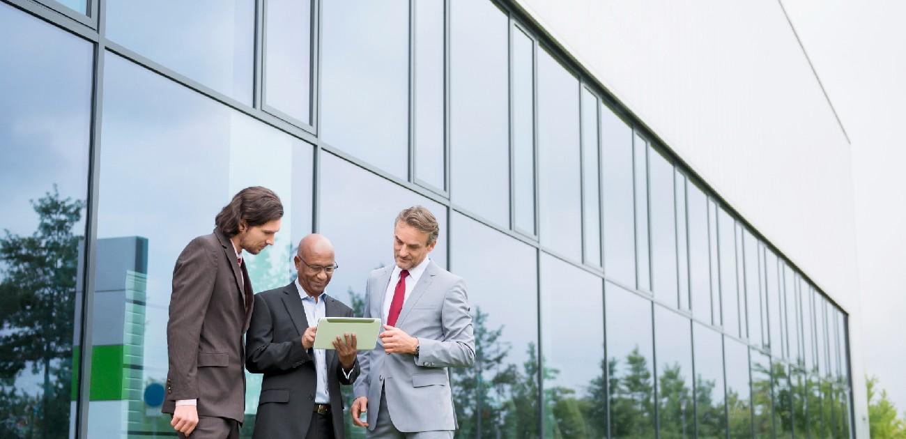 Three people in formal wear stand outside a modern office building all looking at a document together.