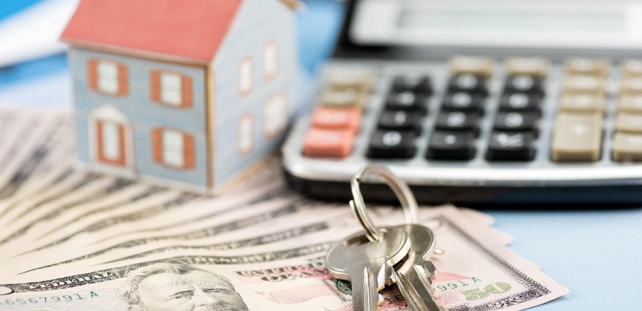 A picture of a set of keys, a house miniature, a calculator, and fanned-out $50 bills on a desk.