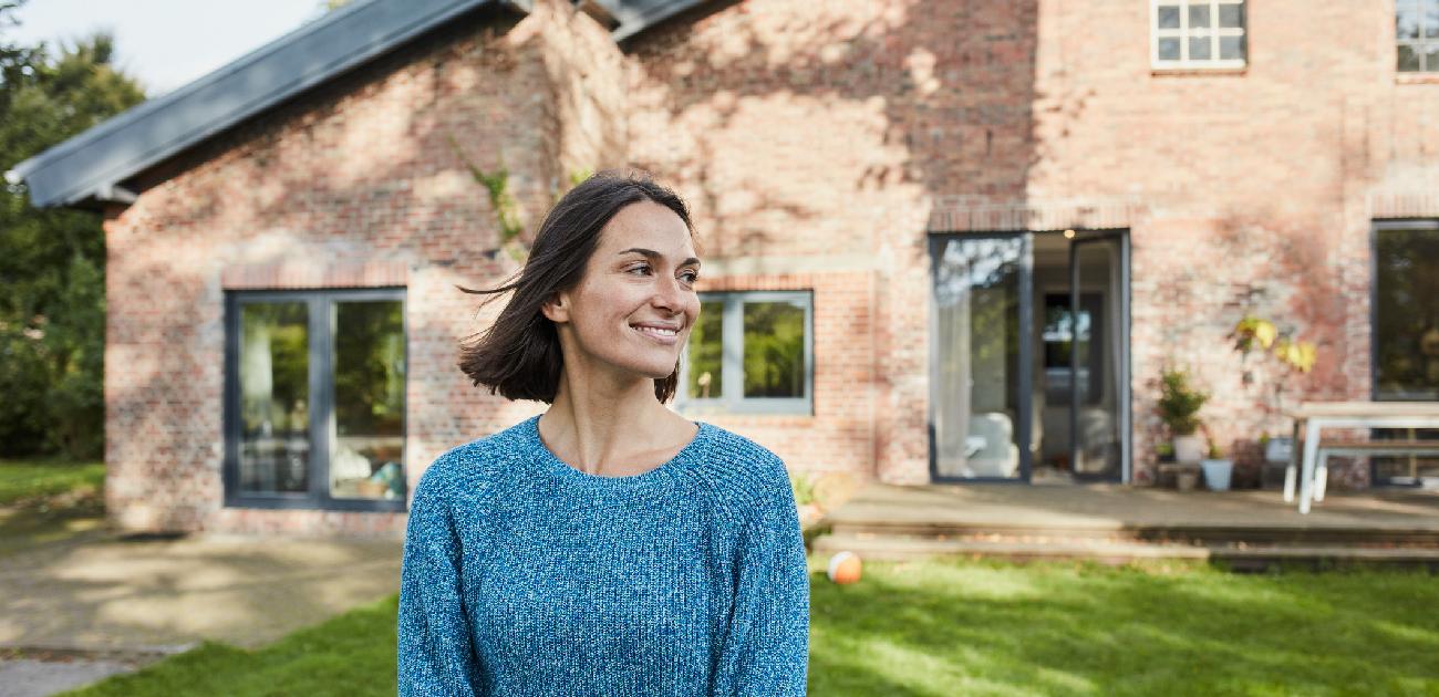 A woman stands in the front yard of a new brick home smiling, looking to her left, right of the frame.