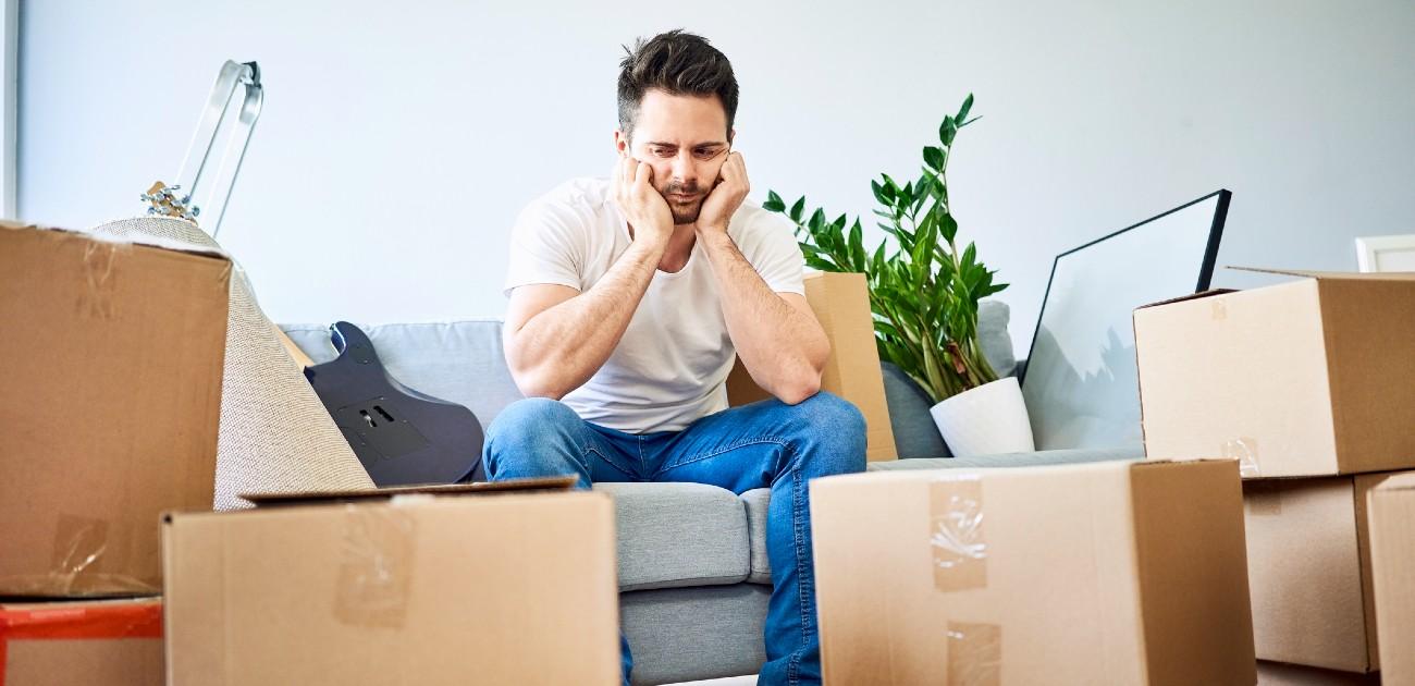 A man sits stressed out with his head in his hands looking at scattered moving boxes.