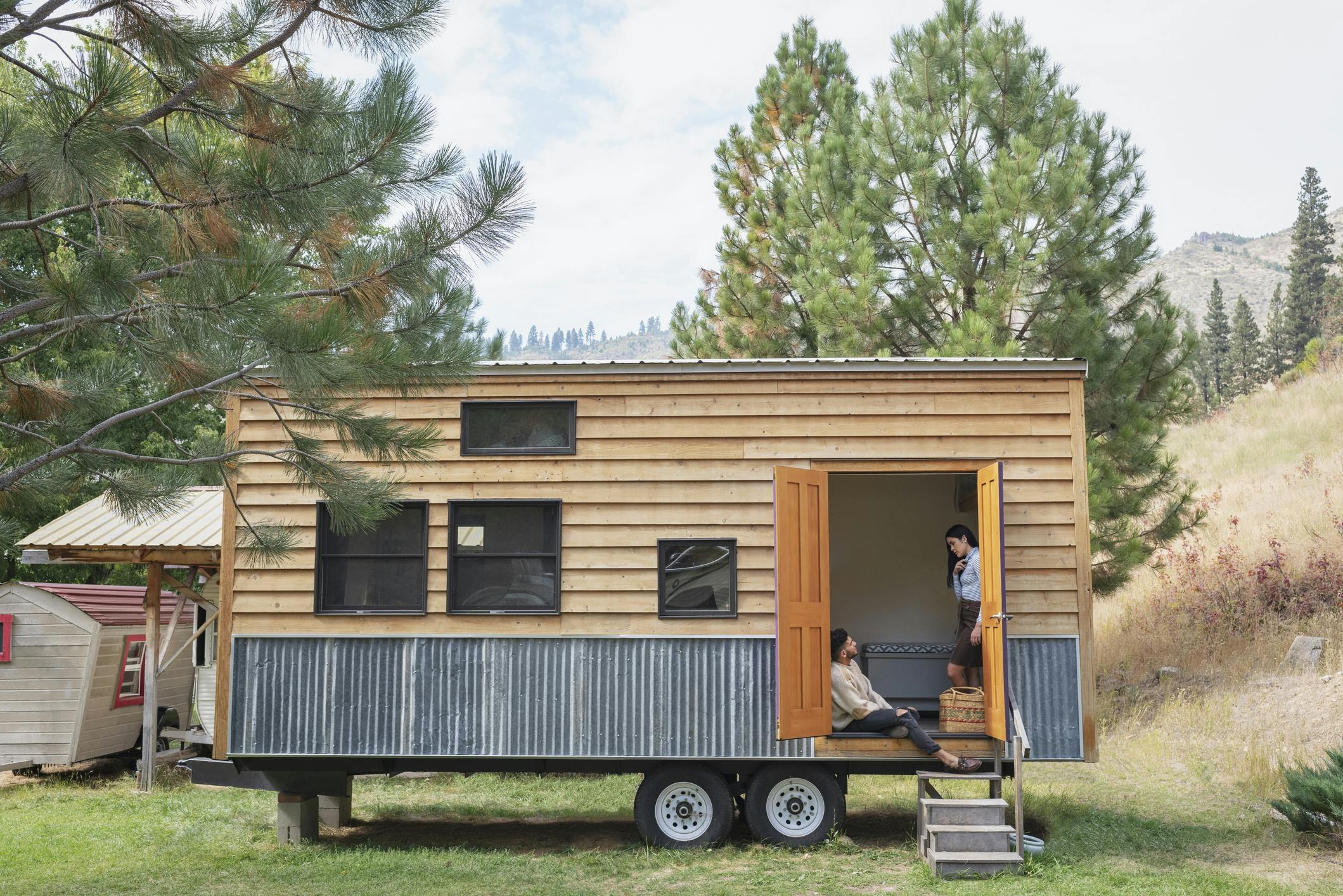 A picture of a tiny home on a wheeled trailer, parked on a grassy lot with hills, trees, and mountains in the background.