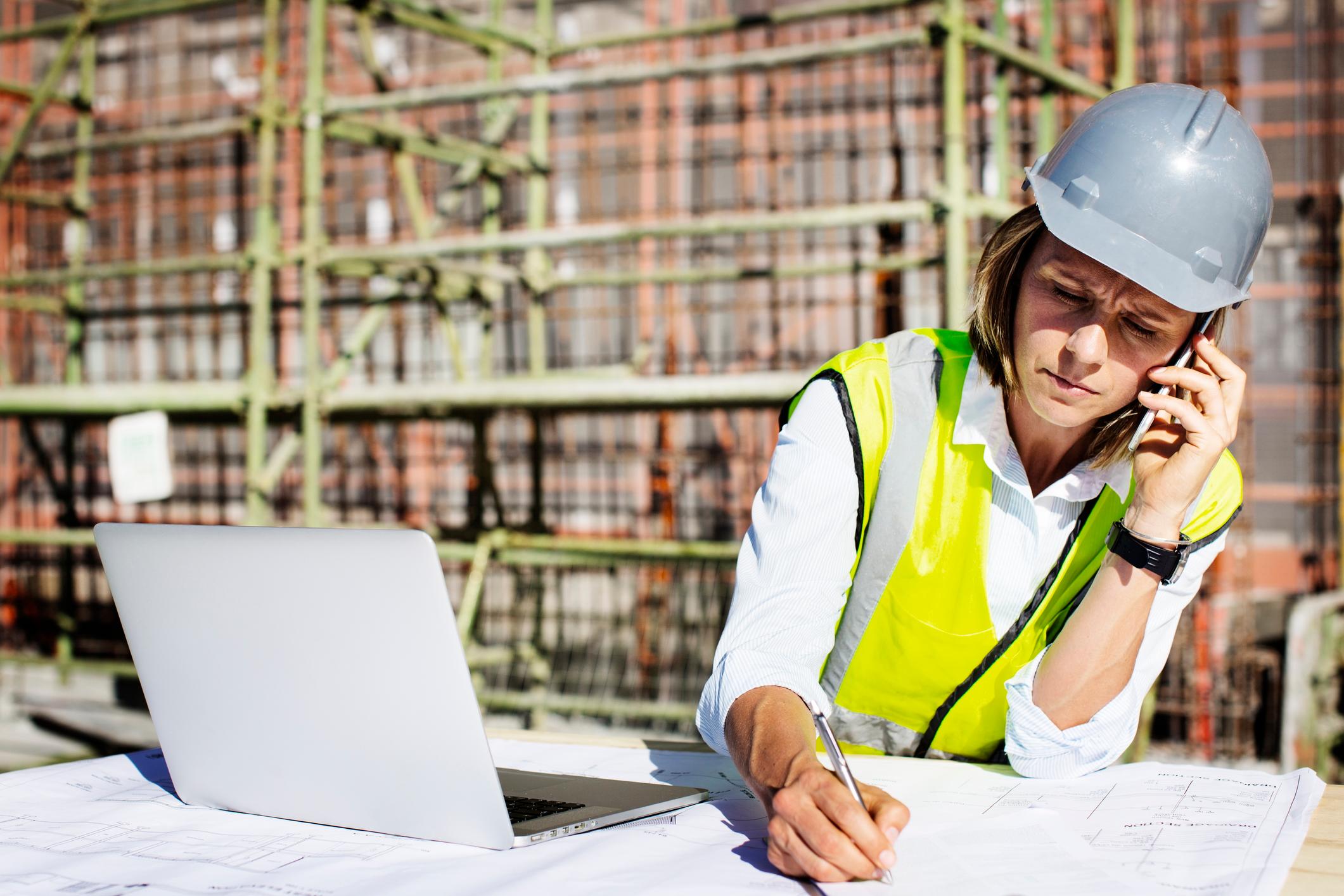 A woman construction worker talking on the phone at a work table with a laptop and building plans she's writing on.