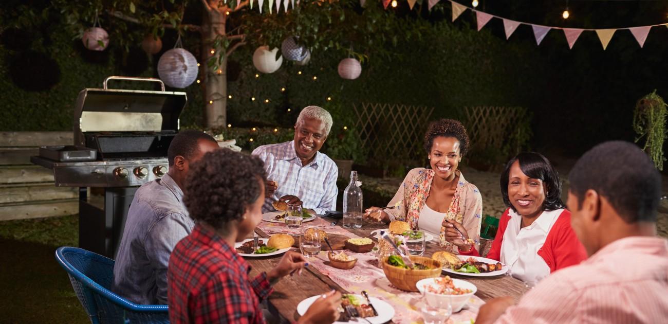 A group of six people sit in a yard at a barbecue enjoying a meal together.