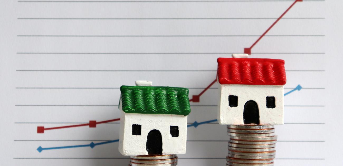 Two house miniatures each upon a stack of coins, one taller than the other with a line graph behind them indicating rising rates