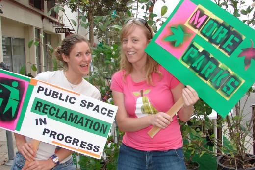 Two women holding up signs that read: "Public Space Reclamation in Progress" and "More Parks"