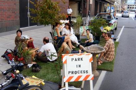 People sitting on the curb behind a sign that reads "Park in Progress"