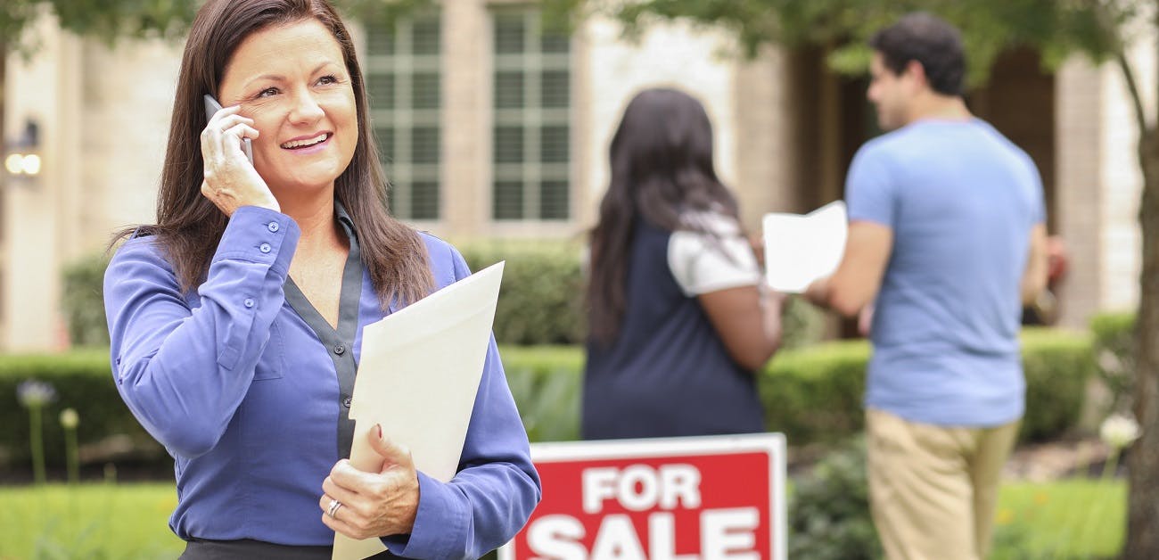 Real estate agent talking on phone with young couple and for sale sign in background