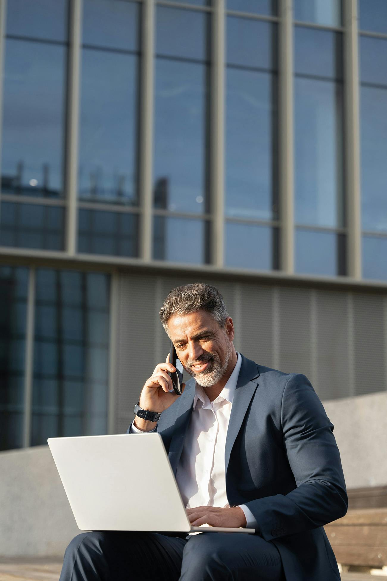 Man in a suit sitting outdoors with laptop talking on cell phone