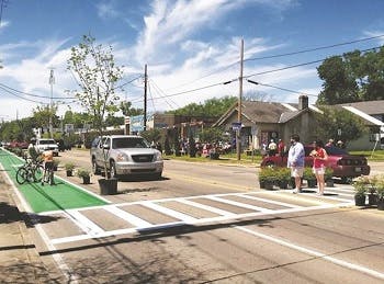 A street scene in a residential area showing people on a green bike lane lined up with plant pots and people crossing the street