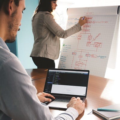 Man working on laptop and woman in background writing notes on a white board