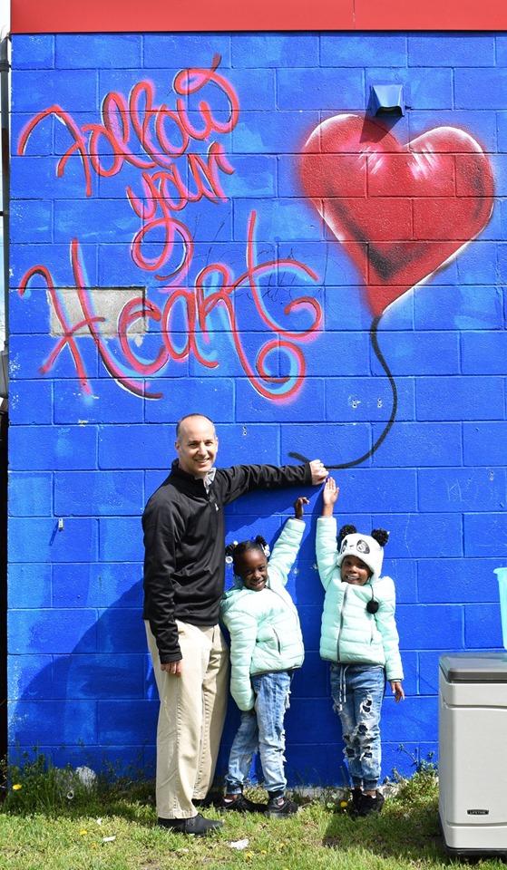 Lansing, MI, Pop-Up Park - man with kids standing in front of a blue wall with a printed message saying follow your heart.