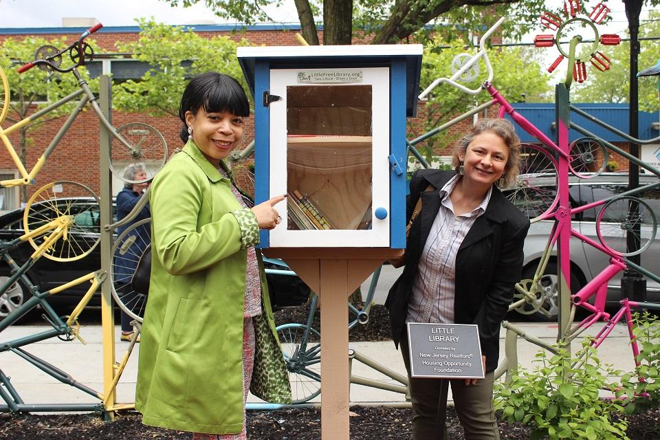 Two women standing right next to the little library and a plaque stating the little library was donated by the NJ REALTORS.