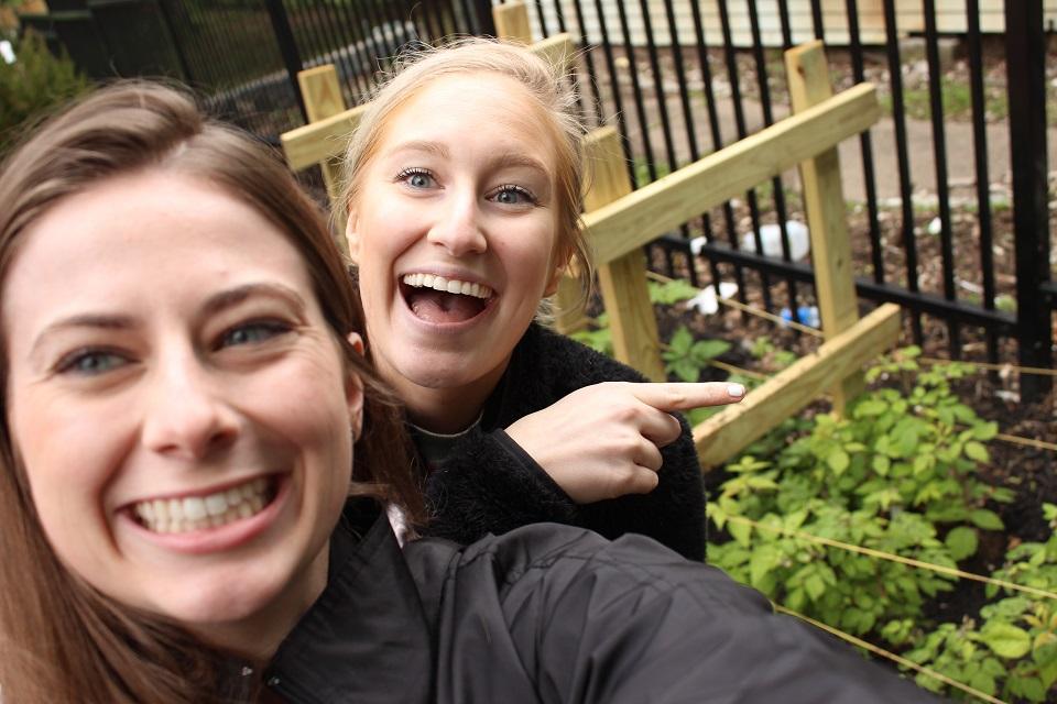 A selfie featuring two women smiling pointing to a gardening bed in the back.