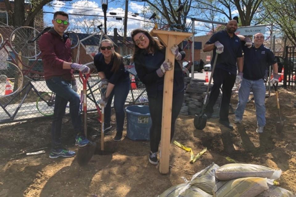 Four people with shovels and one woman holding a piece of wood next to a few sand bags.