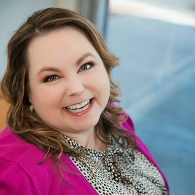 Headshot image of Deanne Rymarowicz in a pink jacket with a blue background.