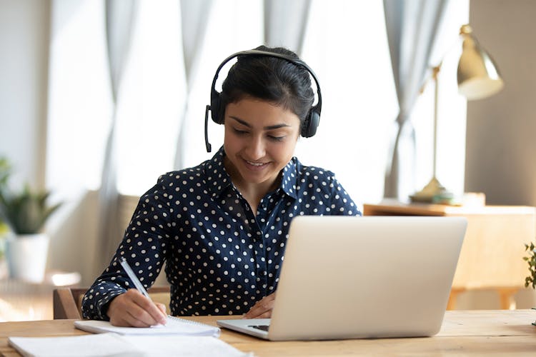 Woman at a laptop, wearing headphones and taking notes